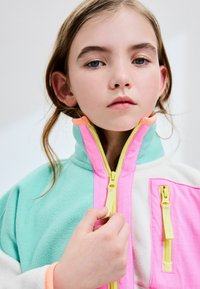 Veste en polaire colorée avec un design vert menthe, rose et crème. Elle comporte plusieurs zips avec des tirettes jaunes et un col haut.