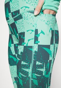 Leggings de sport ajustés en couleur vert teal avec des motifs abstraits en vert foncé. Comprend une poche latérale et des détails de couture visibles.