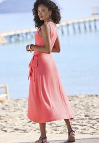 Koralo spalvos be rankovių ilga suknelė su surištu liemeniu, laisvai krentantis audinys ir patogus siluetas, papildytas rudomis sandalais su dirželiais ir paplūdimio aplinka.