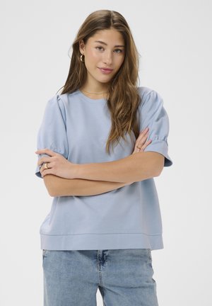 Mladá žena s dlhými hnedými vlasmi v svetlomodrej blúzke s nafúknutými rukávmi a rifľových nohaviciach, so založenými rukami, neutrálnym pozadím.