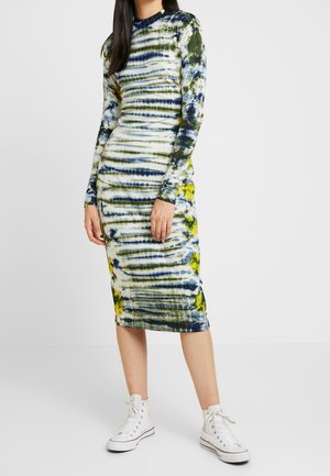 Frau trägt ein enganliegendes, langärmliges, knielanges Batik-Kleid in Weiß, Grün, Blau und Gelb, kombiniert mit weißen High-Top-Sneakers.