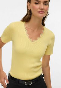 Geel geribd truitje met korte mouwen en een V-hals met geschulpte kanten rand. Draagt een zwarte riem met een ronde metalen gesp.