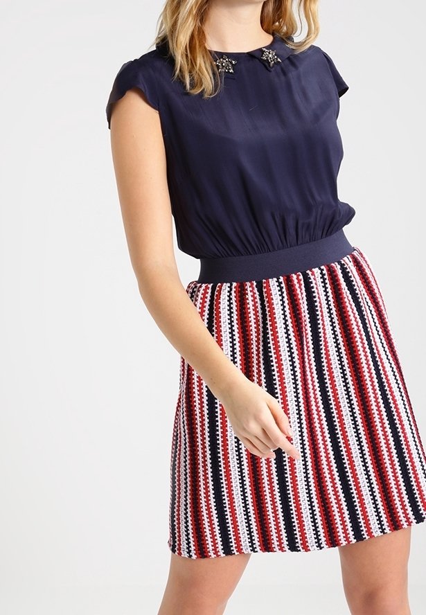 Femme portant un haut sans manches bleu marine avec un col orné et une jupe taille haute à rayures verticales rouges, blanches et noires.