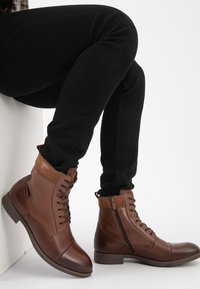 Bruine leren enkellaarzen met suède accenten, vetersluiting aan de voorkant, ritssluiting aan de zijkant en ronde neus; gecombineerd met zwarte slim-fit jeans.