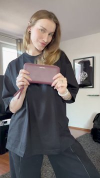 Žena v černém oblečení drží bordó peněženku oběma rukama, stojící v obývacím pokoji s rámovanými uměleckými díly a oknem v pozadí.