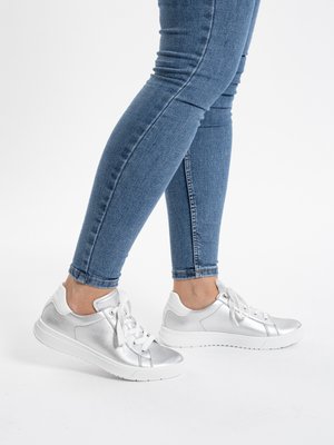 Person wearing blue skinny jeans and silver metallic sneakers with white soles, standing on a white surface.