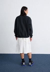 En person står med ryggen til beskueren, iført en sort sweatshirt, hvide shorts og sorte sko med blå accenter, på et blåt tæppe.