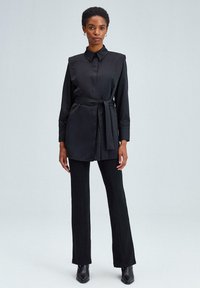 Abito nero camicia con colletto, maniche lunghe e vita cintrata. Texture liscia e satinata, abbinato a pantaloni neri svasati e stivali.