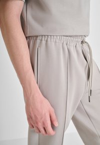 Pantalones jogger de un beige claro con una cintura elástica, detalle de cordón y bolsillos laterales, hechos de una tela suave y lisa.