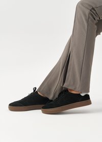 Adidași din piele suede neagră, cu un profil jos, design cu șireturi și talpă din cauciuc maro în contrast, asociați cu pantaloni largi bej.