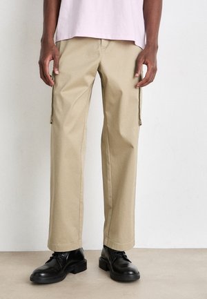 Beżowe spodnie cargo z bocznymi kieszeniami, o prostym kroju nóg i lekkim materiale, połączone z czarnymi sznurowanymi butami.