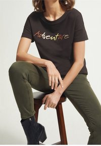 T-shirt nera in cotone con "Adventure" ricamato in multicolore. Abbinarla a jeans verde oliva e stivaletti neri, seduta su uno sgabello di legno.