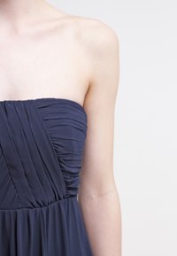 Detailní záběr na osobu v šatech bez ramínek v námořnické modré barvě s plisovanými detaily na korzetu, na jednobarevném pozadí.