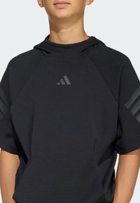 Person som bär en svart kortärmad hoodie med en liten Adidas-logga på bröstet och tre ränder på varje ärm.