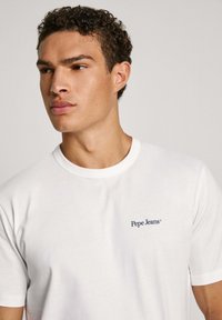 Bílý bavlněný tričko s kulatým výstřihem a krátkým rukávem, s modrým logem "Pepe Jeans" na levé části hrudi. Hladký povrch.