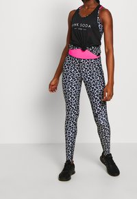 Černý top bez rukávů s logem "PINK SODA", růžový pásek a vzorované černé legíny s geometrickým bílým designem. Černé sportovní boty.