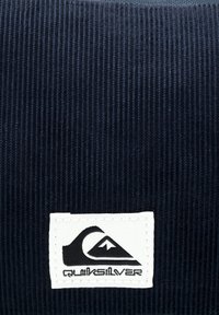 Tmavomodrý kordový materiál má biely obdĺžnikový nášivok s čiernym logom Quiksilver, ktoré znázorňuje vlny a hory.