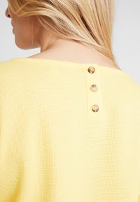 Blondýna v texturovaném žlutém topu se třemi knoflíky na horní části zad, otočená zády k fotoaparátu.