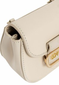 Beżowa skórzana torebka z zaokrągloną klapą, złotymi akcentami i widocznym detalem szwów. Kompaktowy kształt, gładka faktura i minimalistyczny design.