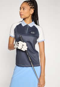 Tumši zila un gaiši zila golfa polokrekls ar strukturētu dizainu, īsām piedurknēm un EAZ logotipu. Modelis tur golfa nūju, uz rokas ir balta cimdiņš.