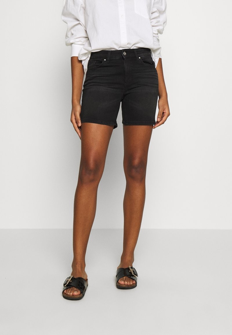 Pantalones cortos de mezclilla negra con un diseño ajustado, cintura de talle medio, cinco bolsillos y dobladillos enrollados, combinados con sandalias planas negras.