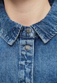 Nærbilde av kragen på en blå denimjakke med to synlige metallknapper, båret av en person med synlig nakkehud.