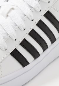 White sneaker with black leather stripes, textured fabric laces, padded tongue, and white rubber sole. Prominent stitching detail.