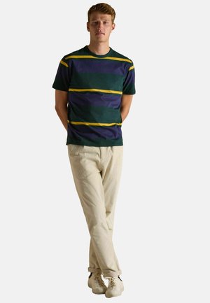 Man standing with hands behind back, wearing a green, purple, and yellow striped T-shirt, beige pants, and white sneakers.