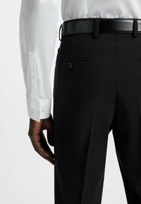 Pantaloni neri su misura con una texture liscia, dotati di una tasca posteriore con bottone e una cintura in pelle nera lucida. Visibile il polsino di una camicia bianca.