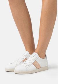 Valged nahast jooksujalatsid beeži suede riba ja perforatsioonide detailidega. Lame talla ja nööpide disain. Suede materjali tekstuur.