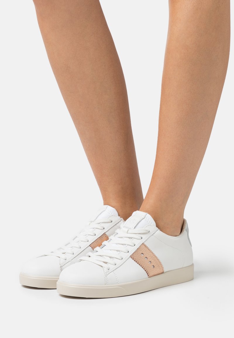 Valged nahast jooksujalatsid beeži suede riba ja perforatsioonide detailidega. Lame talla ja nööpide disain. Suede materjali tekstuur.