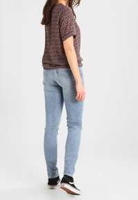 Top de manga corta con estampado en colores oscuros, combinado con jeans ajustados azul claro y zapatillas negras de caña baja, de pie contra un fondo neutro.