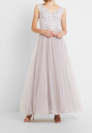 Frau in einem ärmellosen Lavendelkleid mit floraler Spitzenoberseite und Tüllrock, stehend mit gefalteten Händen, trägt offene Absätze.