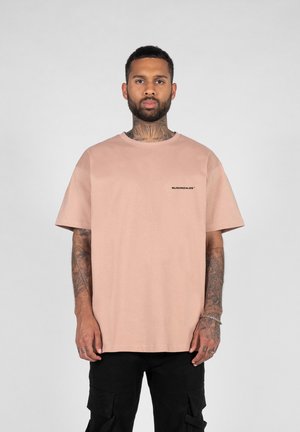 Light pink short-sleeve t-shirt with a crew neck and a small black logo on the chest, made of a smooth fabric. Worn with black pants.