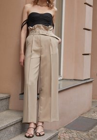 Beige kalhoty s vysokým pasem a širokými nohavicemi s páskem v pase, spárované s černým topem bez ramínek s texturovaným vzorem. Viditelné černé sandály na tenkých popruzích.