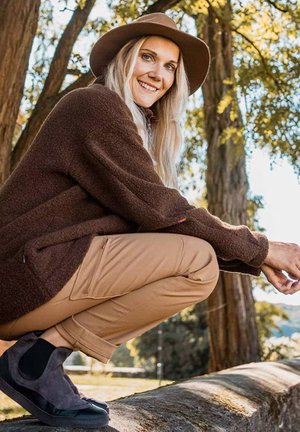 Femme blonde portant un chapeau marron, un pull et un pantalon beige, accroupie sur un rebord en pierre dans un parc ensoleillé avec de grands arbres.