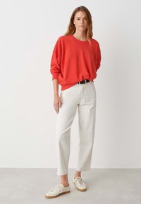 Sweatshirt rouge avec des épaules tombantes, en tissu doux, associé à un pantalon droit blanc et des baskets beiges, sur un fond clair.