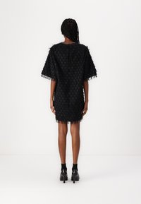 Robe noire avec un motif texturé, manches courtes, détails de frange à l'ourlet et coupe droite. Portée avec des chaussures à talons hauts noires.
