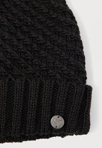 Μαύρο πλεκτό σκουφί με ριγωτό σφράγισμα και μεταλλικό λογότυπο. Η υφή είναι απαλή, με ορατό παχύ νήμα και αδιάσπαστο σχέδιο.