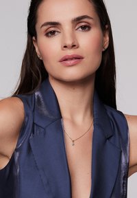 Granatowa satynowa marynarka z klapami i błyszczącą fakturą. Nosi ona delikatny złoty naszyjnik z małym niebieskim wisiorkiem.