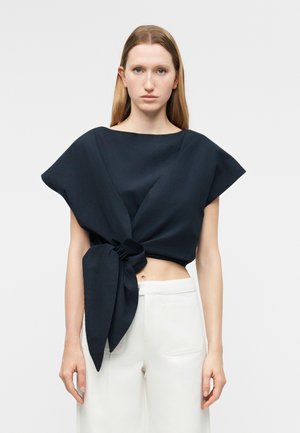 Mujer con una blusa negra asimétrica sin mangas, anudada en la cintura, combinada con pantalones blancos de talle alto, de pie contra un fondo liso.