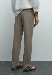 Man in beige broek, wit opgerold lang shirt en zwarte slippers staand op een gespikkelde donkere vloer tegen een effen muur.