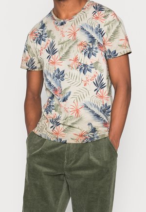 Mężczyzna w beżowej koszulce z krótkim rękawem z wielokolorowym nadrukiem tropikalnych liści i zielonych sztruksowych spodniach, z rękami w kieszeniach, na jednolitym tle.