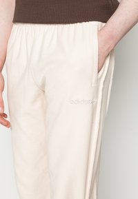 Pantalones de chándal de color crema con una tela texturizada, cinturilla elástica, bolsillos laterales y sutil logo de "adidas" en blanco. Corte relajado.