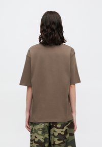 Tricou din bumbac maro cu guler rotund, mâneci scurte și croială lejeră. Împerecheat cu pantaloni cu un model de camuflaj verde.