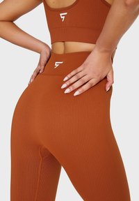 Brązowe legginsy sportowe z prążkowanej tkaniny z wysokim stanem i białym detalem z logo. Tkanina wydaje się elastyczna o gładkiej fakturze.