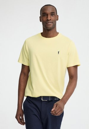 Homme portant un t-shirt jaune pâle à manches courtes avec un petit logo de golfeur sur la poitrine, associé à un pantalon bleu foncé et une ceinture tressée.