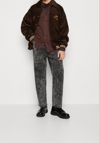 Brun manchesterjacka med randiga ärmar, över en vinröd mönstrad skjorta. Grå slitna jeans och svarta skor fulländar looken.