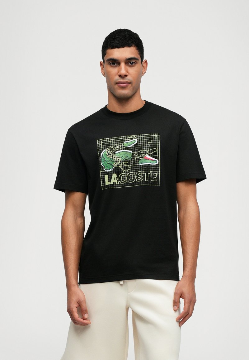 Μαύρη βαμβακερή μπλούζα με κοντό μανίκι που διαθέτει έναν πολύχρωμο γραφικό σχεδιασμό με sneakers και την λέξη "LACOSTE," με στρογγυλή λαιμόκοψη.