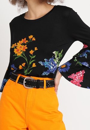 Chemise noire à manches longues avec des imprimés floraux colorés, associée à un pantalon orange vif taille haute et une ceinture noire cloutée.